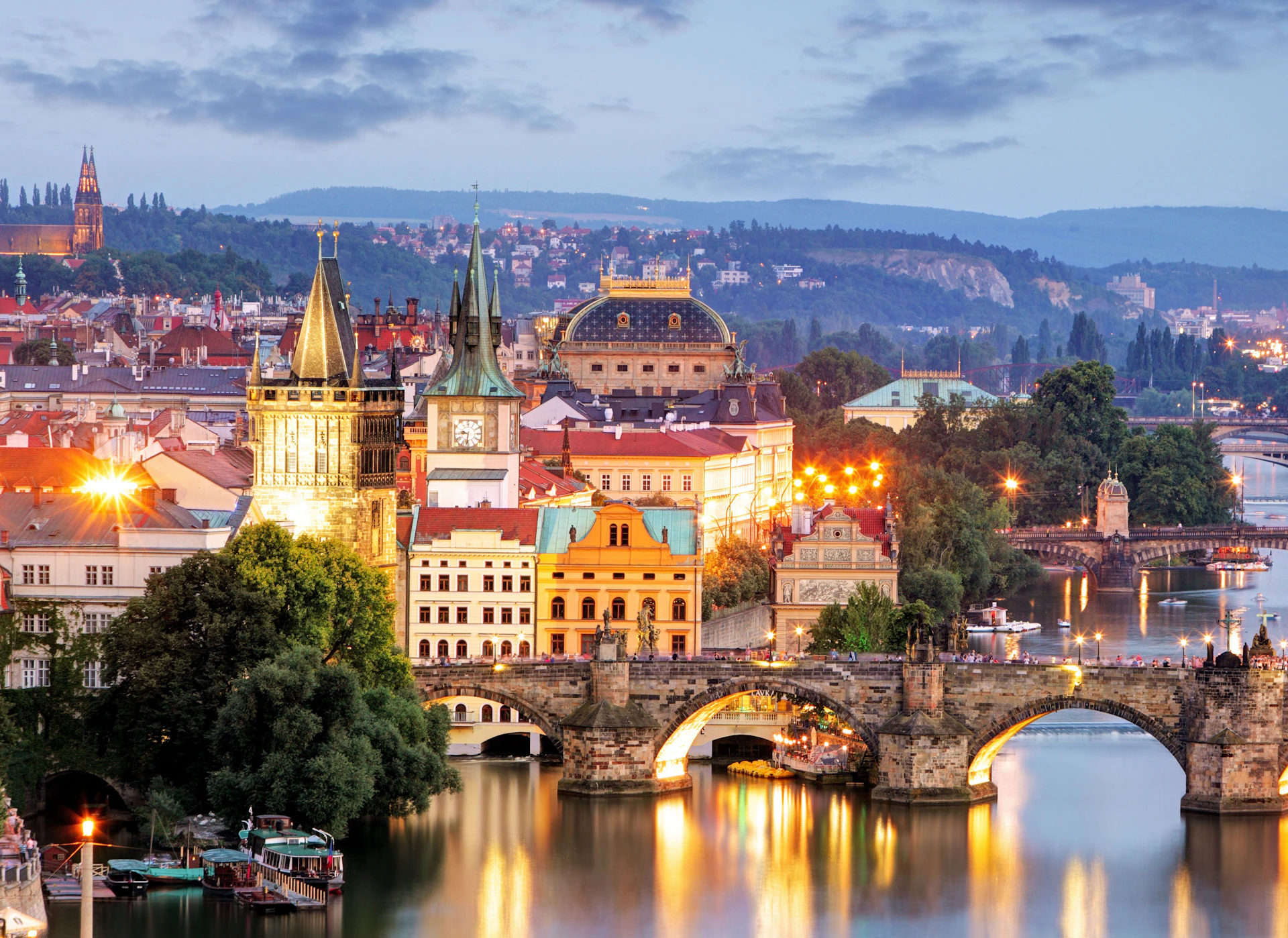 Historic bridge and illuminated buildings over a river, with a backdrop of a cityscape and hills during twilight.