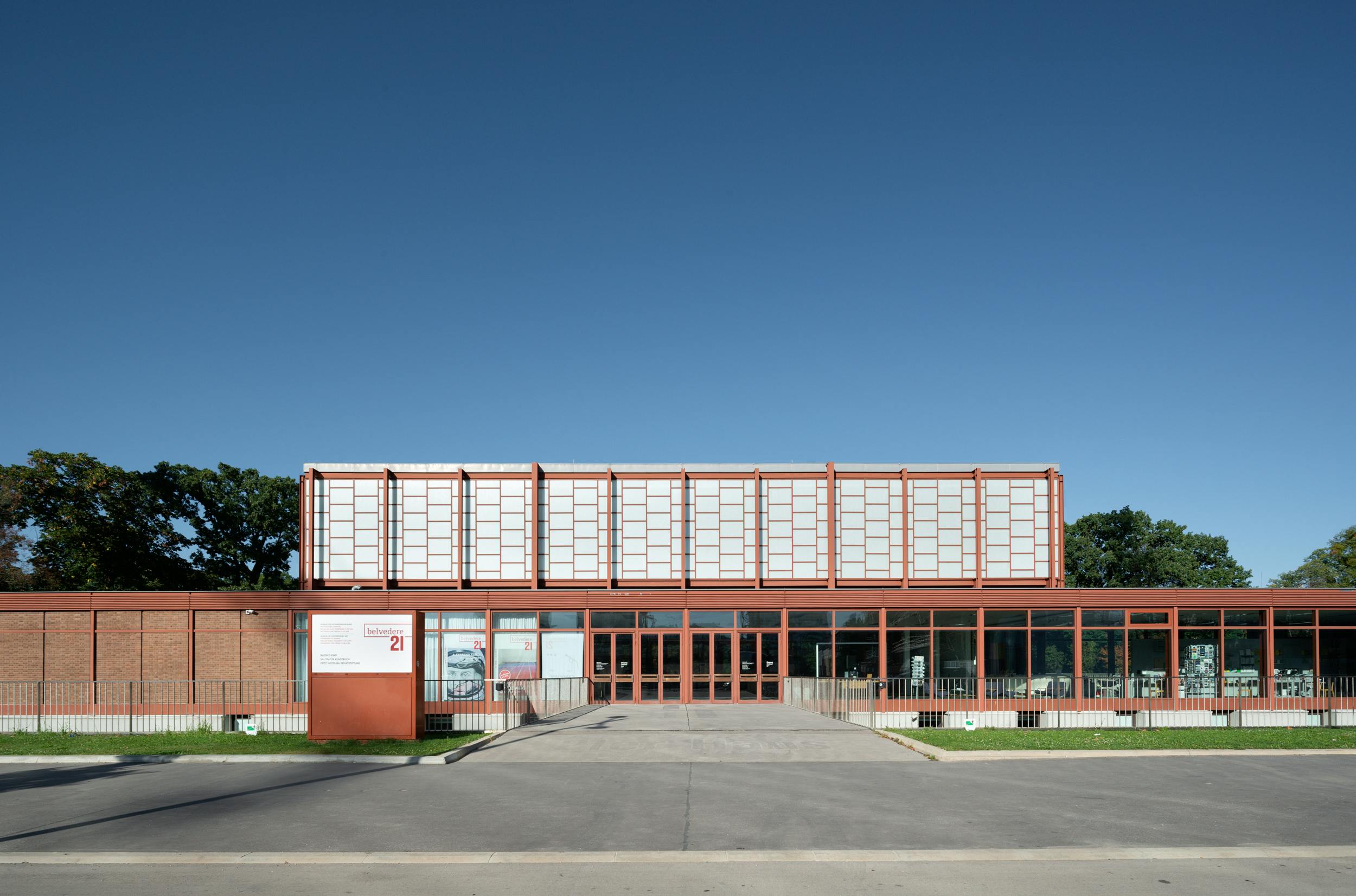 Μοντέρνο κτίριο με μεγάλα παράθυρα και κόκκινο πλαίσιο, με κύρια είσοδο και σήμανση στην αριστερή πλευρά, κάτω από έναν καταγάλανο ουρανό.