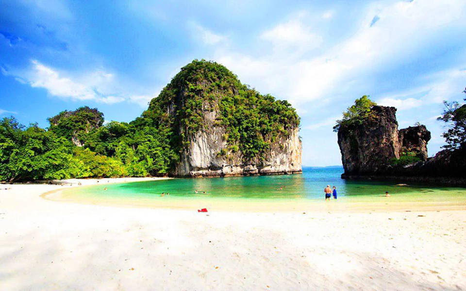 Clear beach with white sand and turquoise water, large rocky cliffs covered in greenery. Few people swimming and relaxing.
