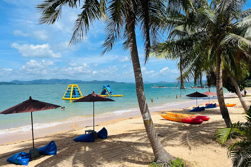 Sandy beach with palm trees, kayaks, and umbrellas. People swimming by inflatable structures in the clear blue ocean.