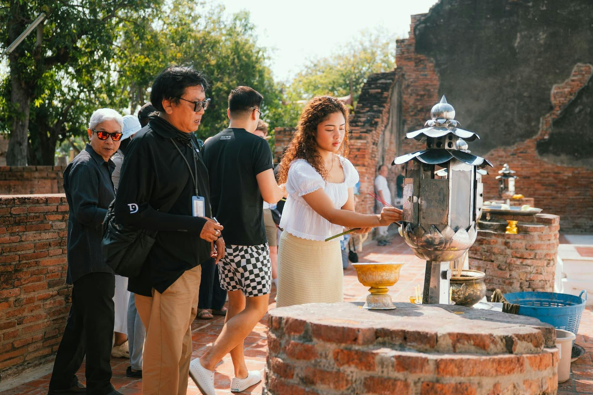 En gruppe mennesker står i nærheden af en murstensstruktur udendørs på en solskinsdag med træer synlige i baggrunden.