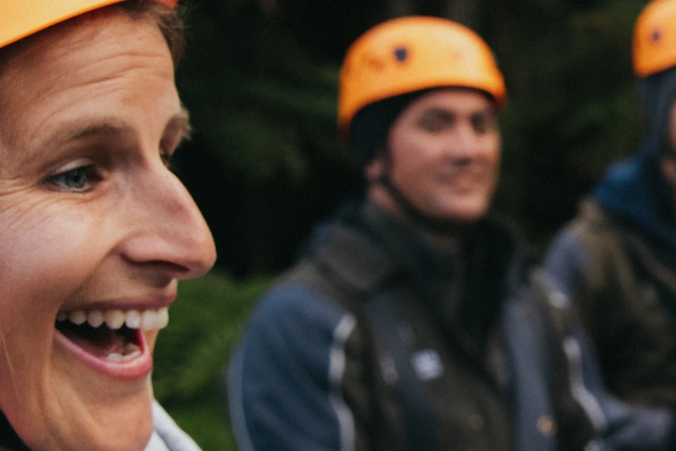 A smiling person wearing an orange helmet holds up a small object, with two others in similar gear in the background outdoors.