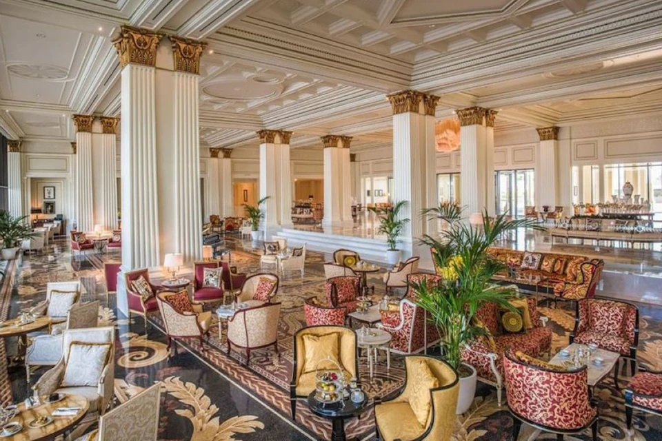 Elegant hotel lounge with ornate columns, patterned carpets, upholstered chairs, potted plants, and chandeliers.