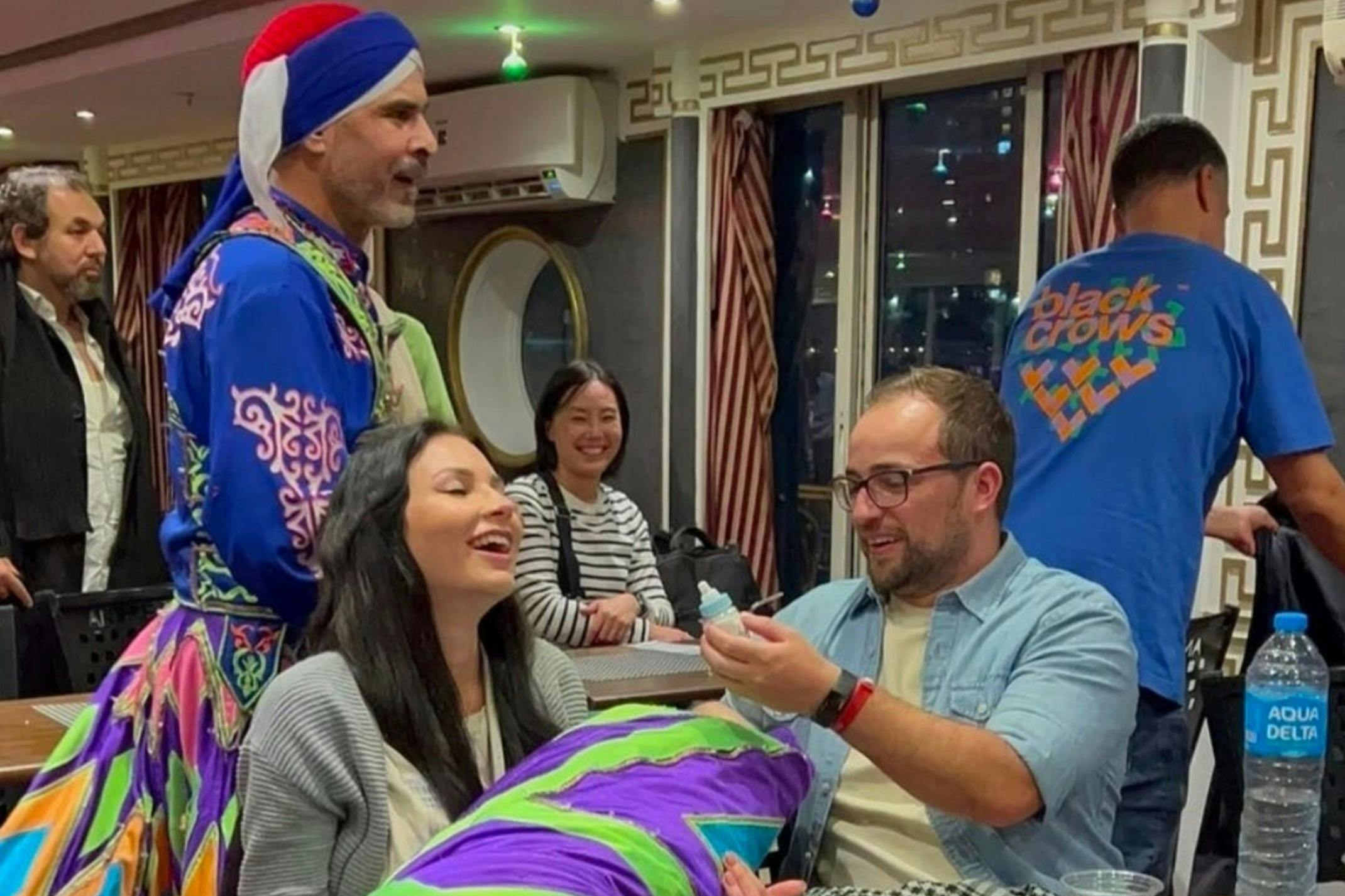 Ein Mann in farbenfroher Tracht tritt in einem Restaurant auf, während die Menschen in der Nähe lachen und den Moment genießen.