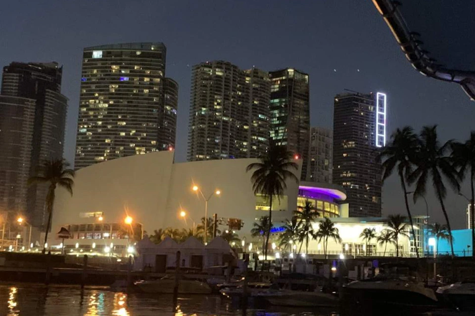 A nighttime cityscape featuring tall buildings, a lit modern structure, palm trees, and a waterfront with boats.