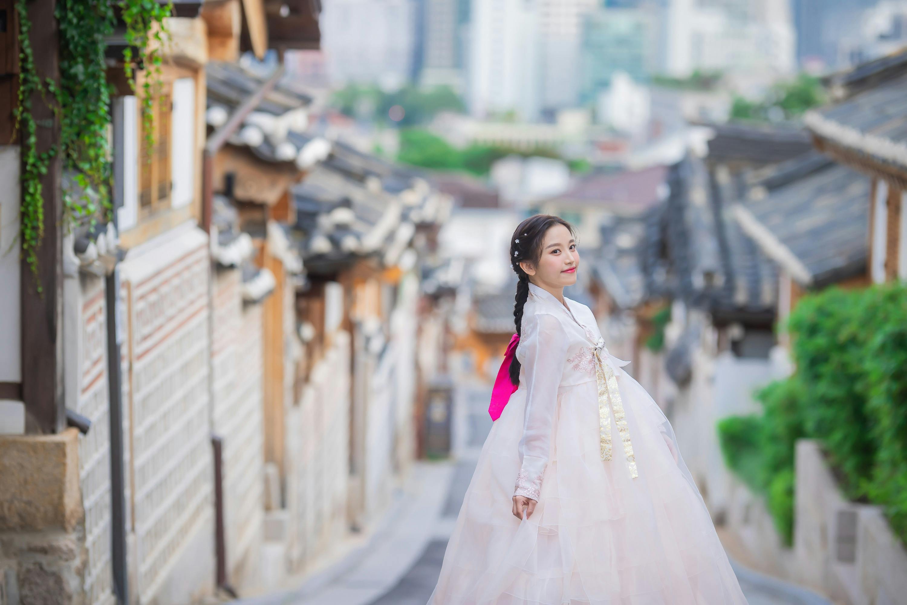 A woman in traditional Korean attire stands on a picturesque street with traditional houses and a cityscape in the background.