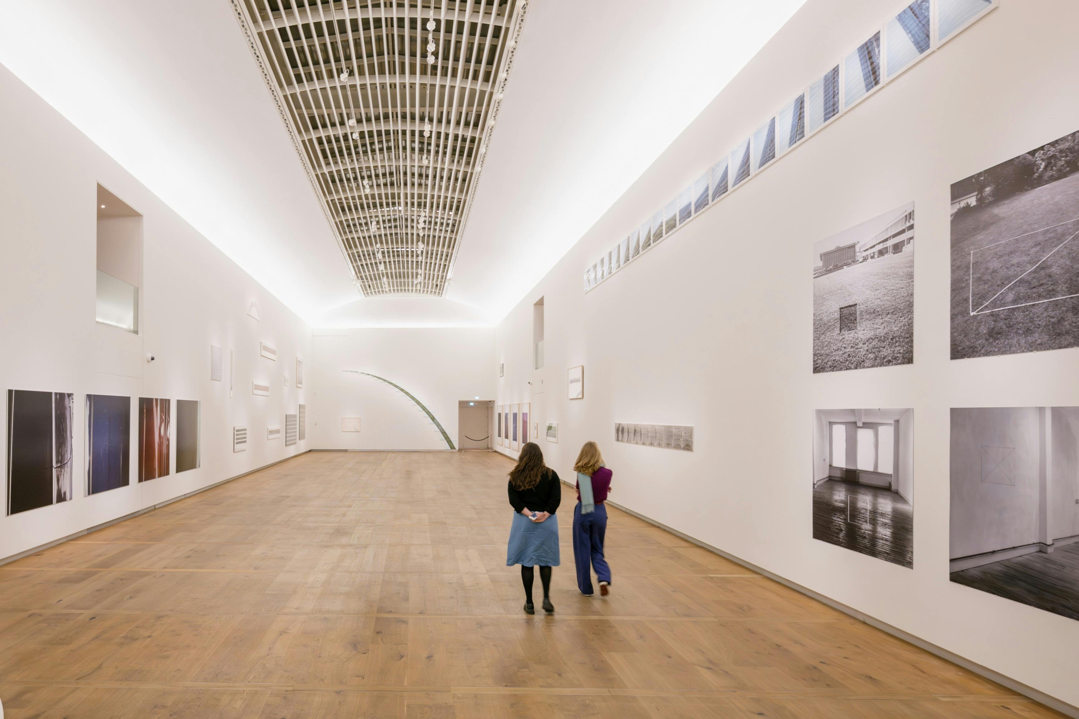 Twee mensen observeren kunstwerken in een moderne galerie met witte muren, houten vloeren en hoge plafonds.