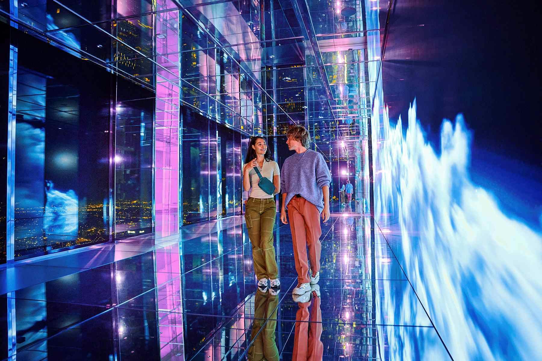 Two people walk through a mirrored tunnel with vibrant pink, purple, and blue lights creating a futuristic atmosphere.
