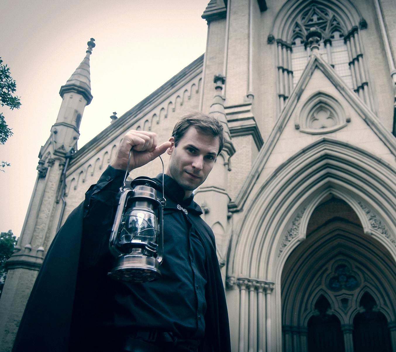 一位男士拿着一盏灯站在一栋哥特式建筑前，这栋建筑配有拱形窗户和华丽的细节。