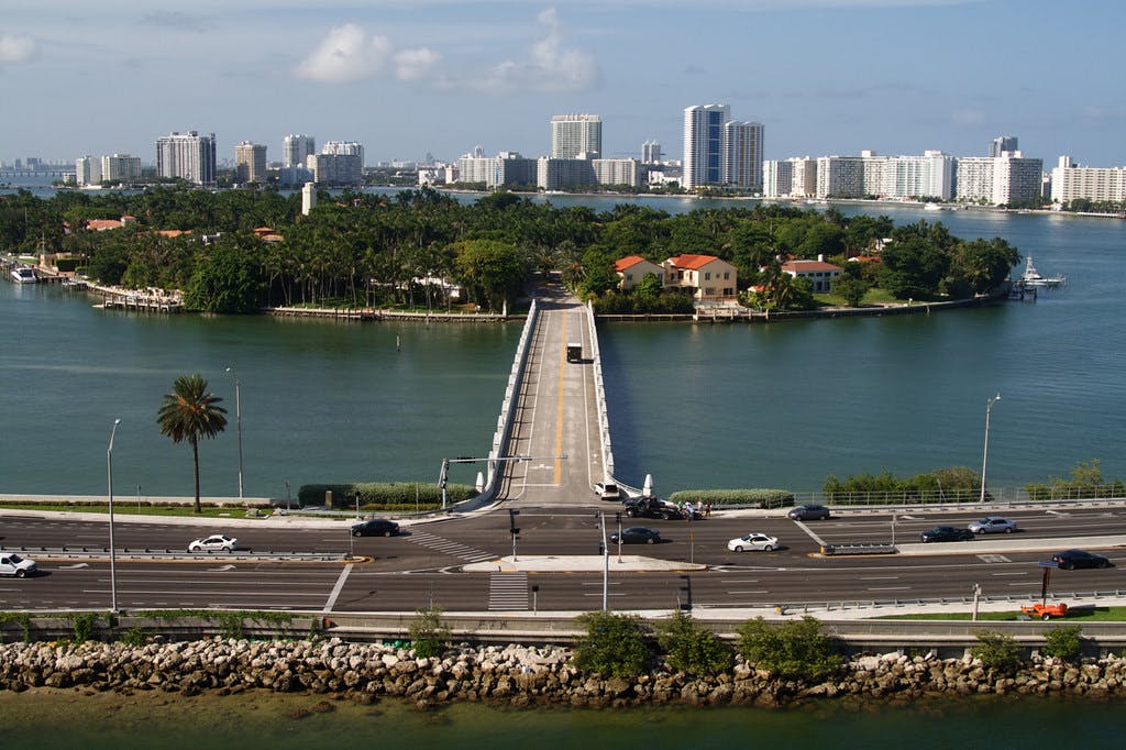 A bridge connects a lush island with red-roofed houses and palm trees to a mainland city with high-rise buildings across a body of water.