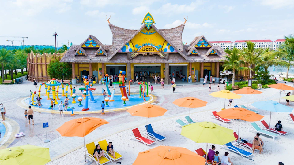 A colorful water park with a central play area, yellow building labeled 'Locker,' and lounge chairs under umbrellas in the foreground.