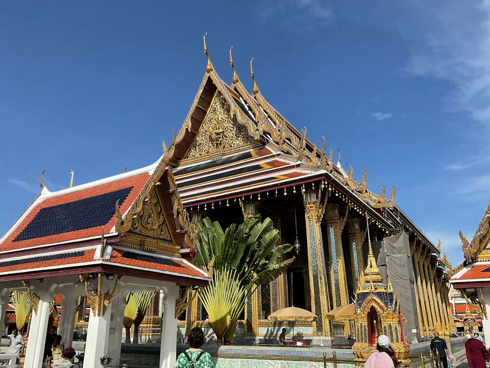 A richly adorned temple with ornate gold details and a sloping roof under a clear blue sky, with tourists in the foreground.