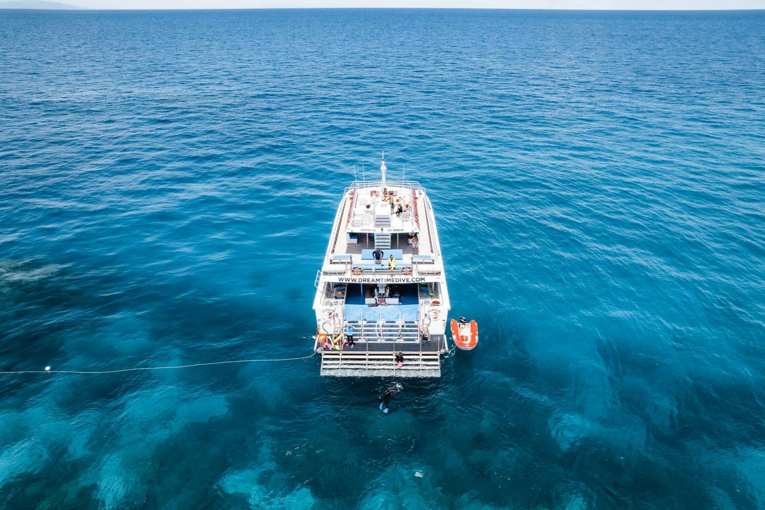 A boat named "Dreamtime Dive" anchored in clear blue ocean water with people on deck and a small dinghy nearby.