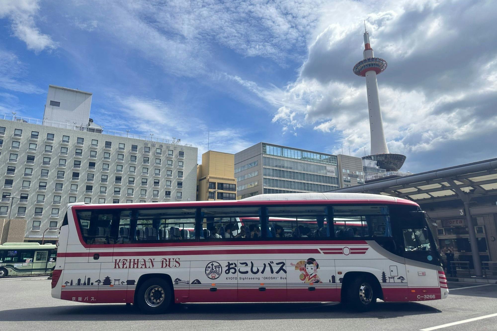 Ένα λεωφορείο για αξιοθέατα σταθμευμένο σε μια αστική περιοχή με τον Πύργο του Κιότο και διάφορα κτίρια στο παρασκήνιο κάτω από έναν εν μέρει συννεφιασμένο ουρανό.