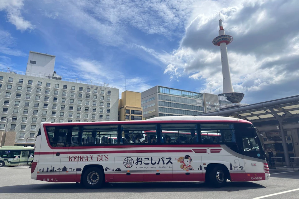 A sightseeing bus parked in an urban area with Kyoto Tower and various buildings in the background under a partly cloudy sky.