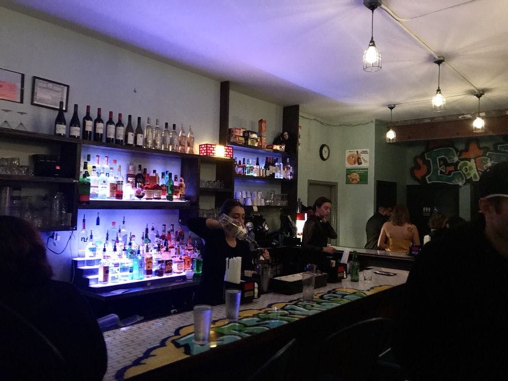 酒吧灯光昏暗，设有涂鸦装饰的柜台，调酒师为您倒饮料，顾客坐在吧台旁边。