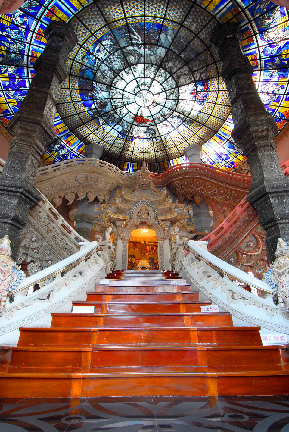 Ornate interior with a grand, richly decorated staircase leading to a vivid stained glass dome, featuring intricate carvings and columns.