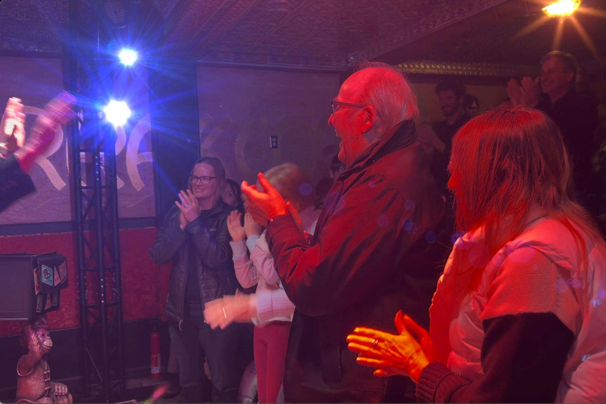 En gruppe mennesker klapper i et svagt oplyst indendørs rum med farverig belysning og dekorationer.