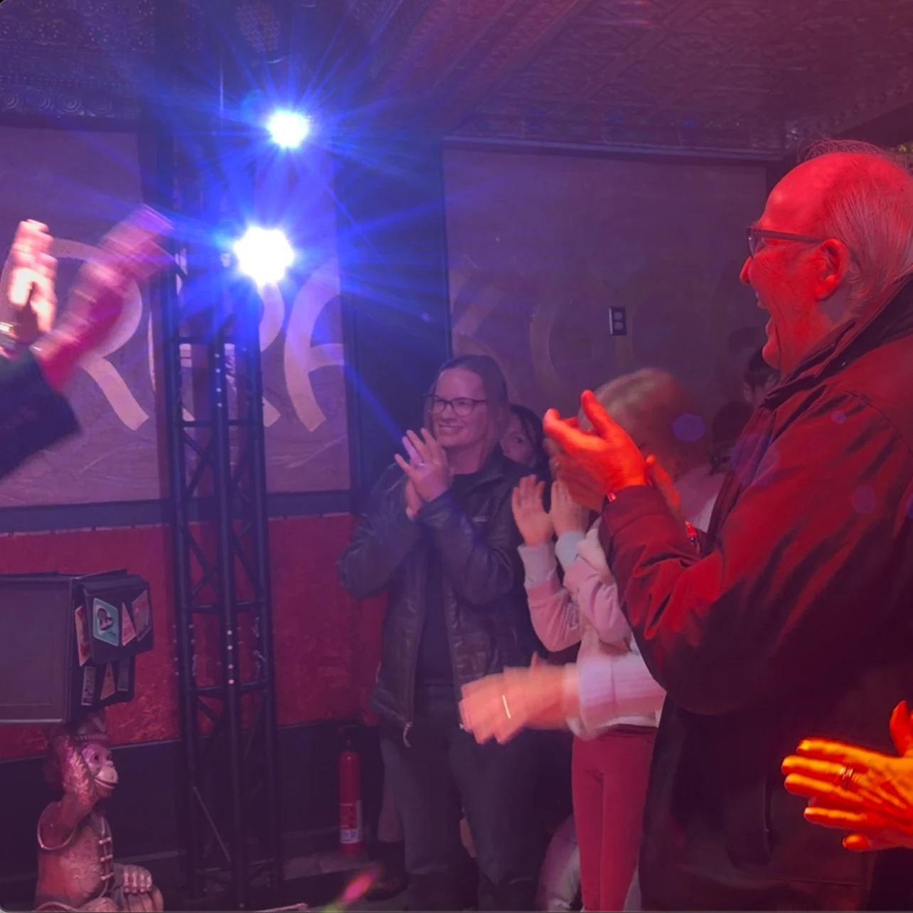 A group of people clapping in a dimly lit indoor space with colorful lighting and decorations.