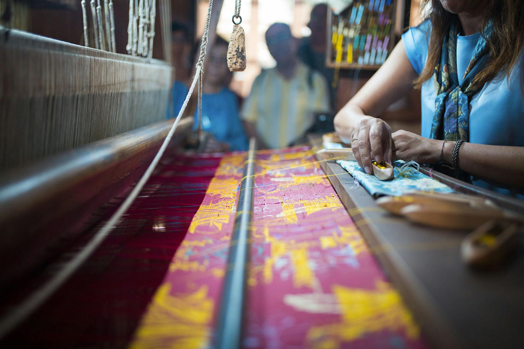 糸やスプールを前面に、織機にカラフルな生地を織っている人と、バックグラウンドで見ている人のクローズアップ。
