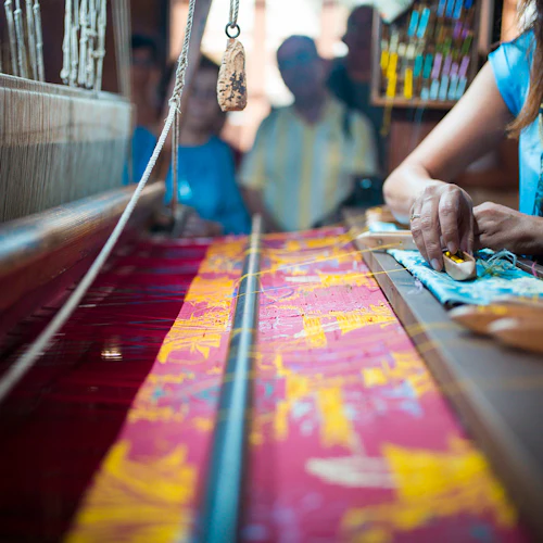 Närbild av en person som väver färgglatt tyg på en vävstol, med trådar och spolar i förgrunden och människor som tittar på i bakgrunden.