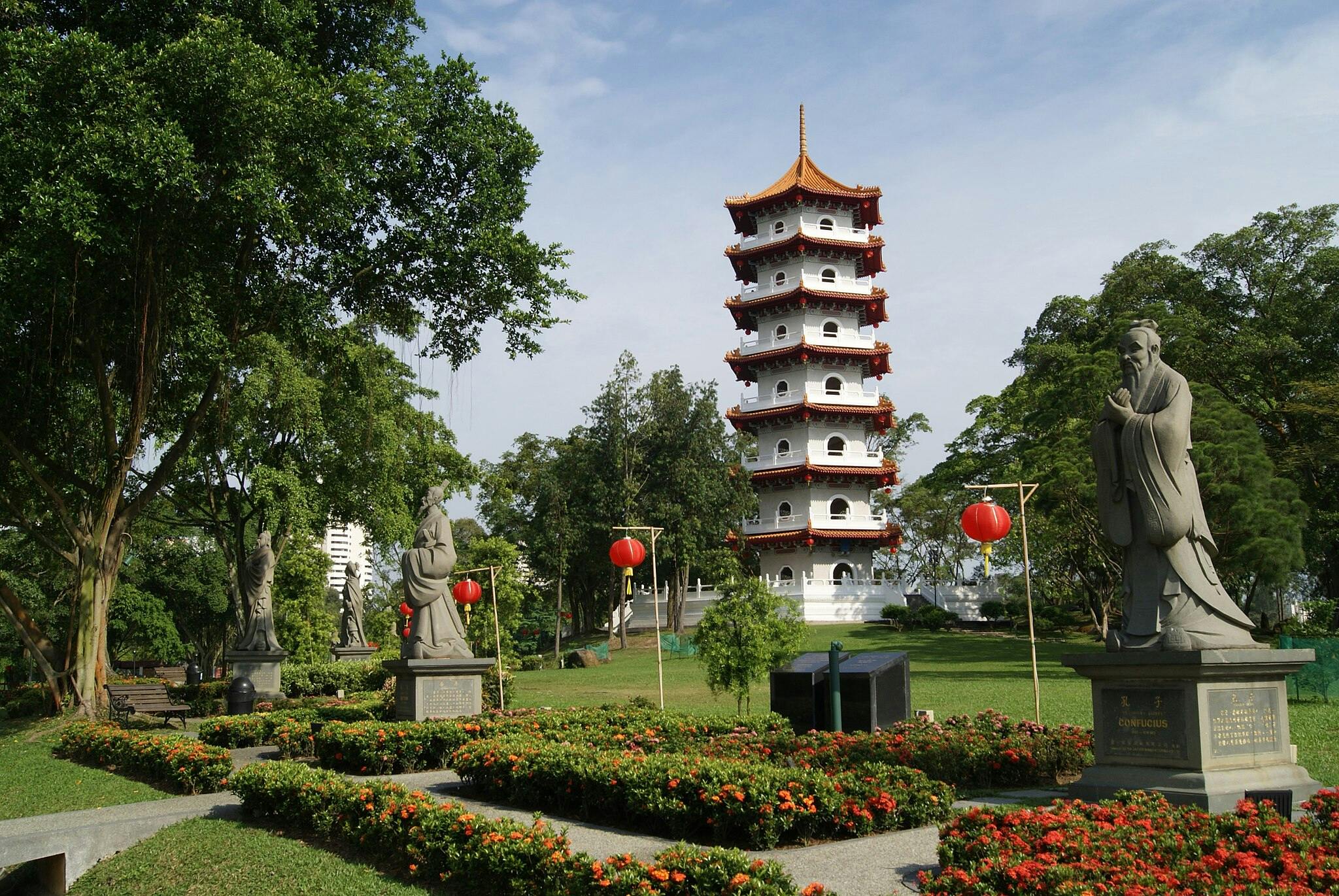 Pagode traditionnelle à étages rouges et blancs dans un jardin, entourée de statues, de lanternes rouges, d'arbres et de fleurs épanouies.