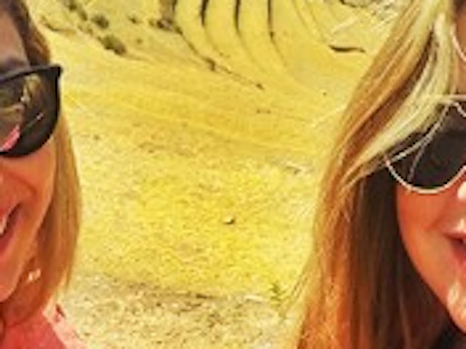 Two women smiling in a sunny mountainous landscape with terraced hills and blue sky with scattered clouds.