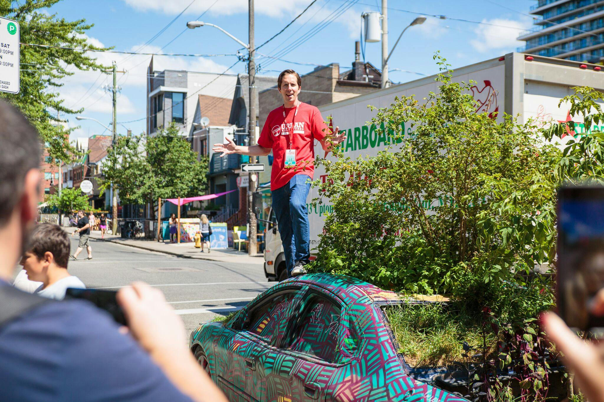 A man wearing a red shirt stands on a colorfully painted car, gesturing, while people nearby take photos on a sunny street.
