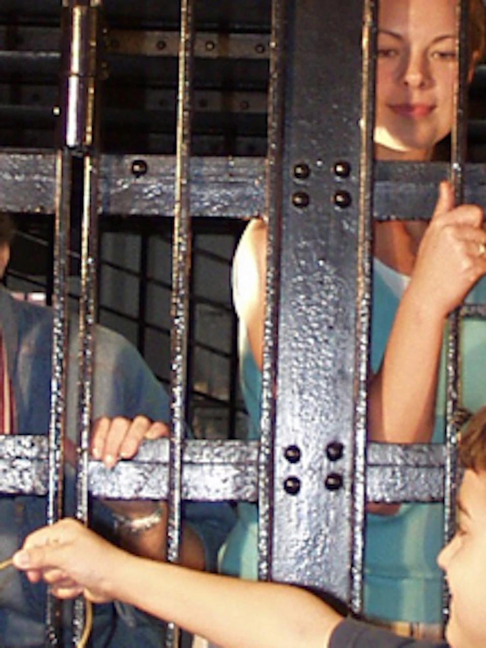 Four adults and a child standing inside a large black jail cell, while two other children stand outside holding keys.