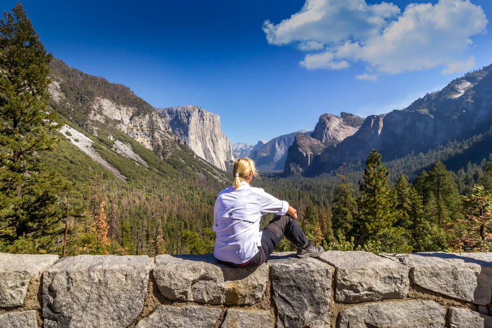 Person sitting on a stone wall, overlooking a valley with mountains and forests under a clear blue sky with scattered clouds.
