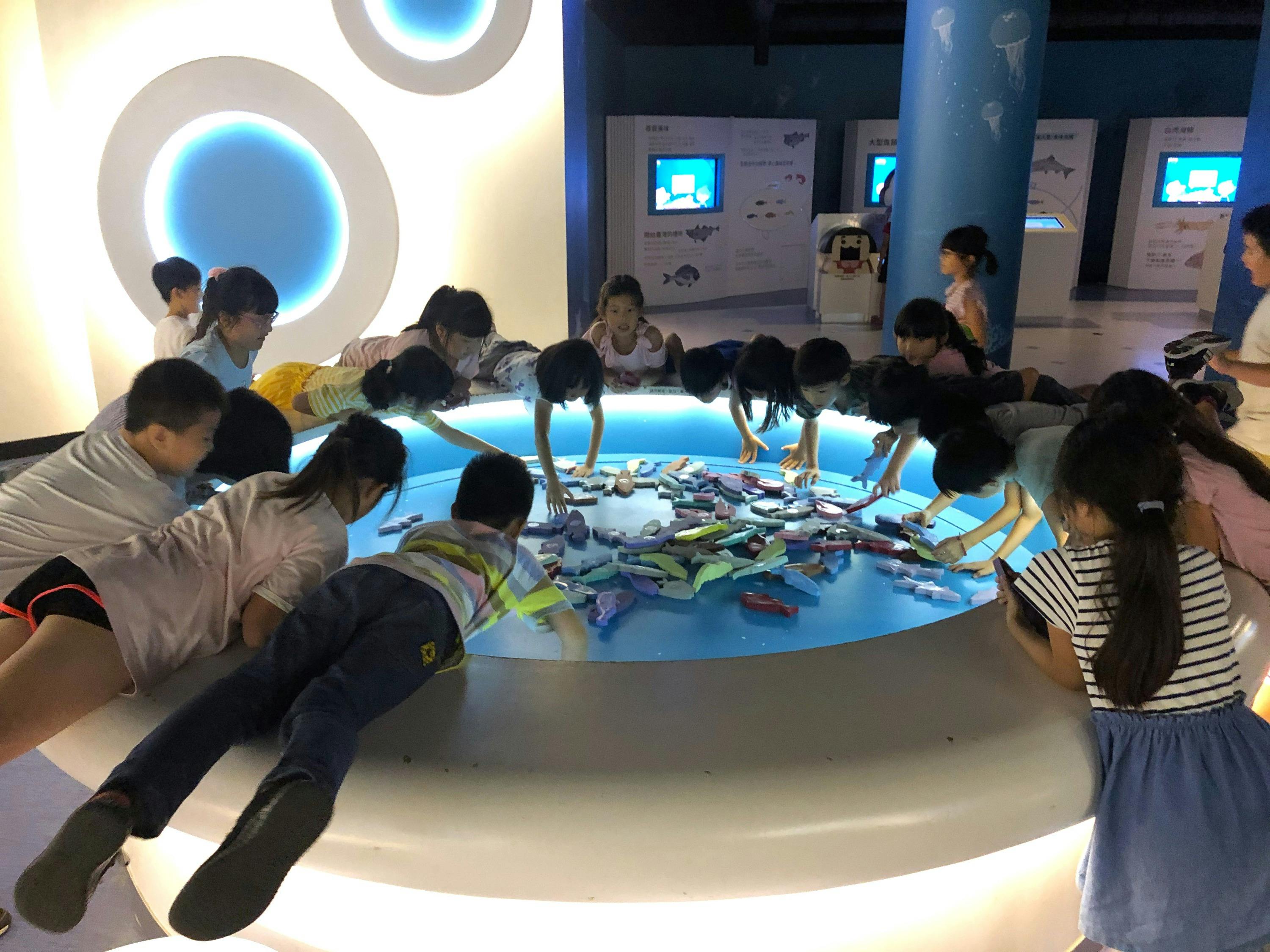 Children leaning over a glowing circular display table in a brightly lit, modern exhibition space.
