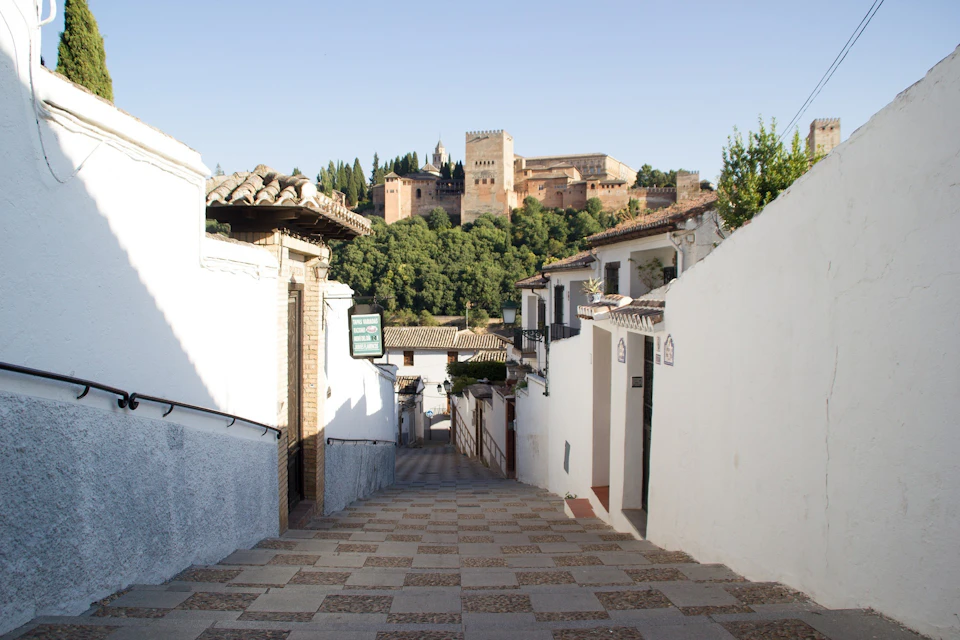 Narrow cobblestone street flanked by white buildings, leading to a distant castle surrounded by trees under a clear sky.