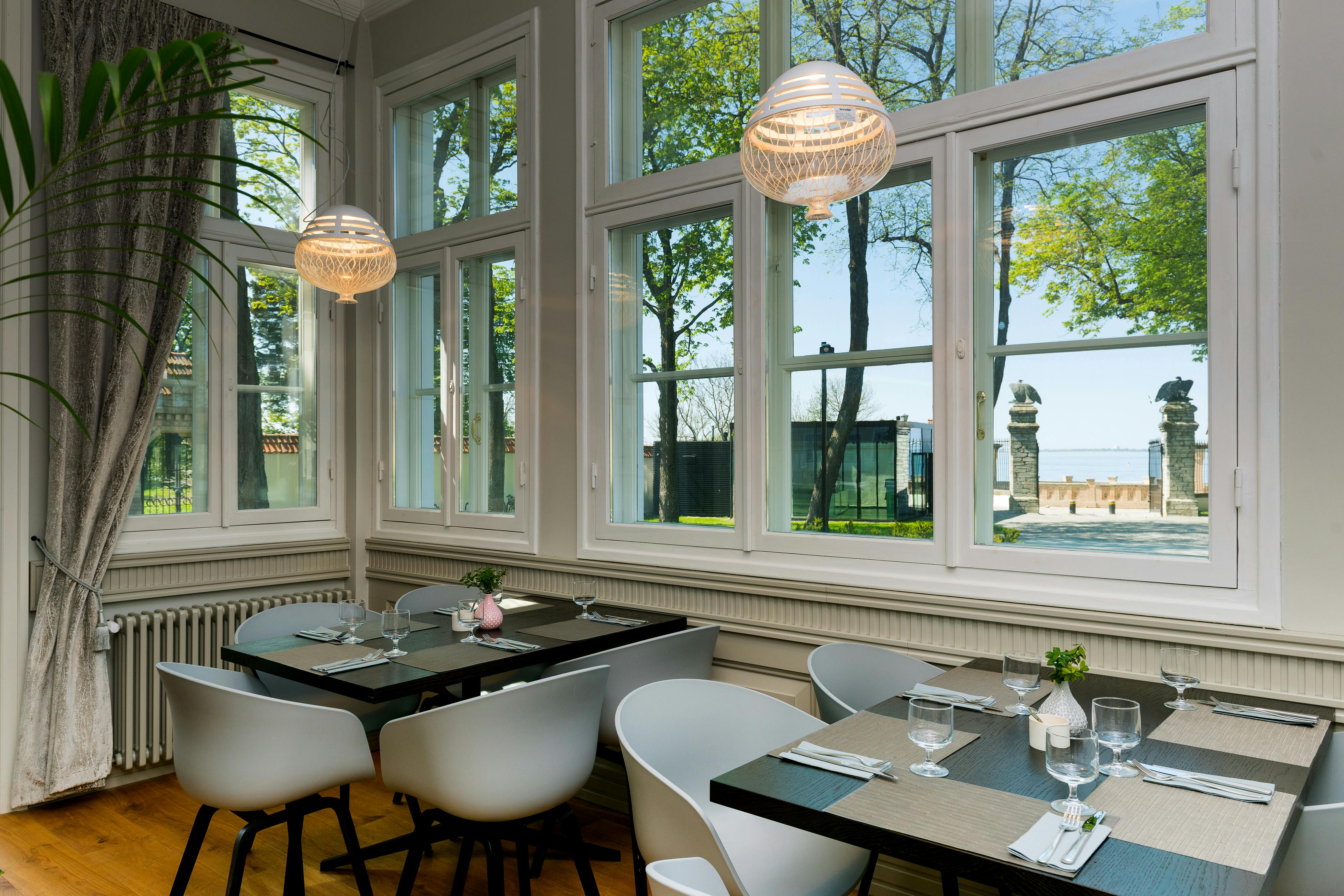 Sala de jantar iluminada pelo sol com janelas, cadeiras brancas, mesas escuras com copos e talheres, candeeiros suspensos e árvores visíveis no exterior.