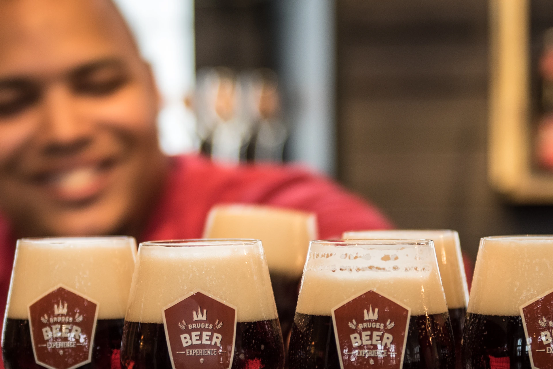 A smiling bartender in a red shirt stands behind a tray of six foamy dark beers on a counter.