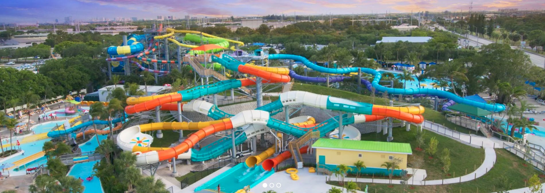 Large water park with numerous colorful water slides, pools, and a few people enjoying the attractions among trees and buildings.