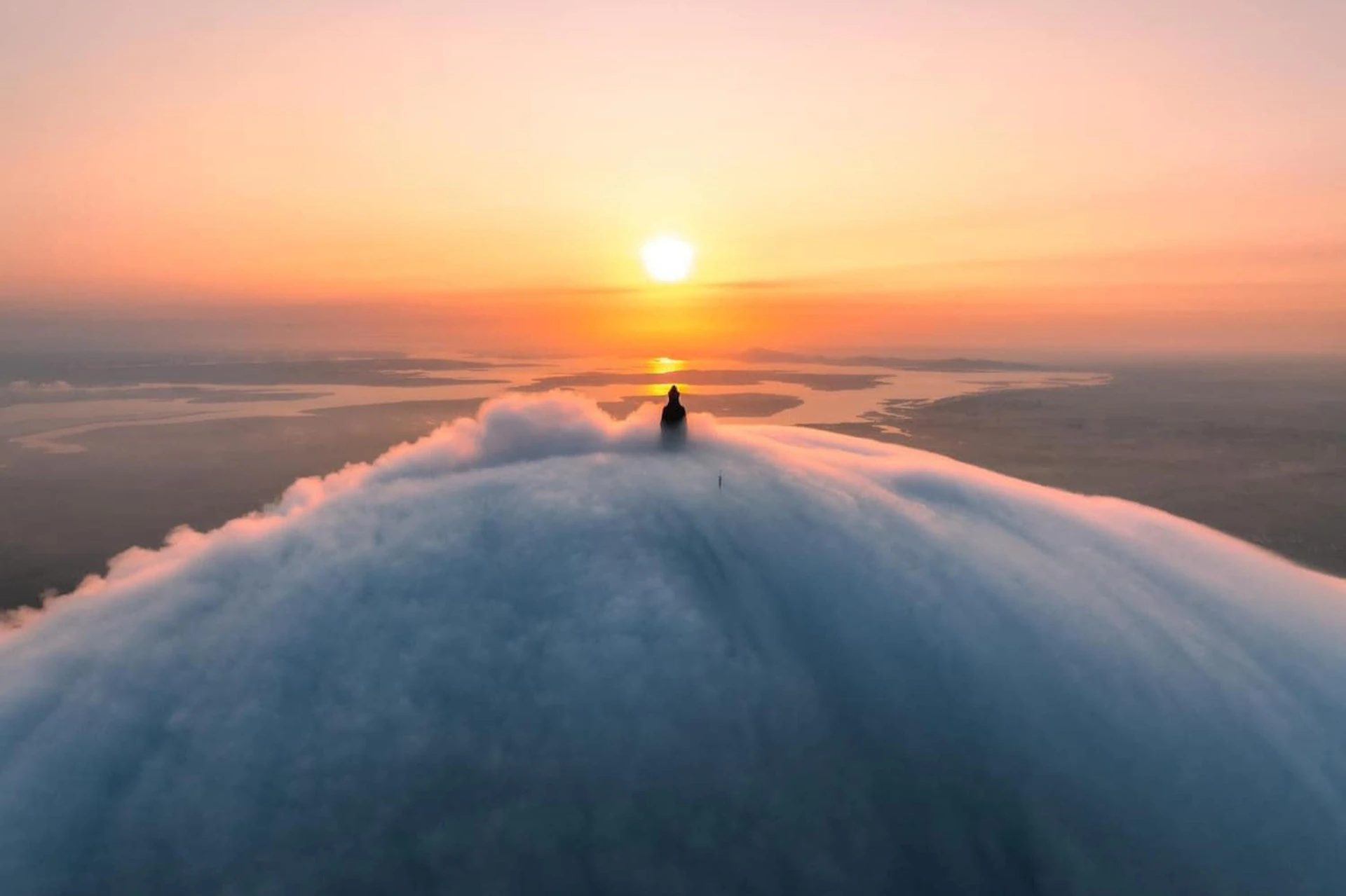 A person stands on a cloud-surrounded peak, facing a distant sunset over a calm, expansive landscape.