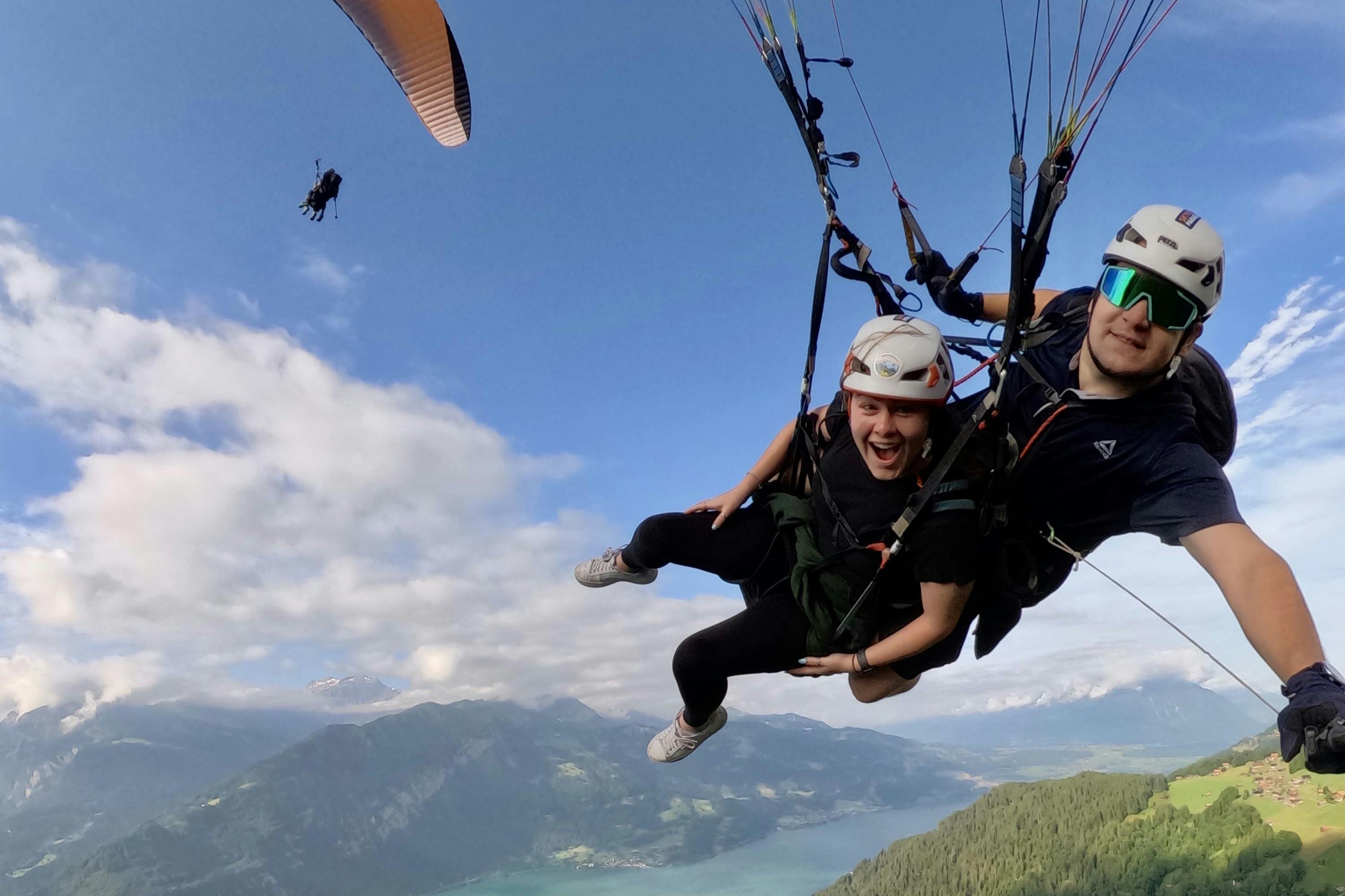 Deux personnes font du parapente avec des montagnes, un lac et une forêt en contrebas. L'une des personnes sourit largement. Les deux personnes portent des casques et des harnais.
