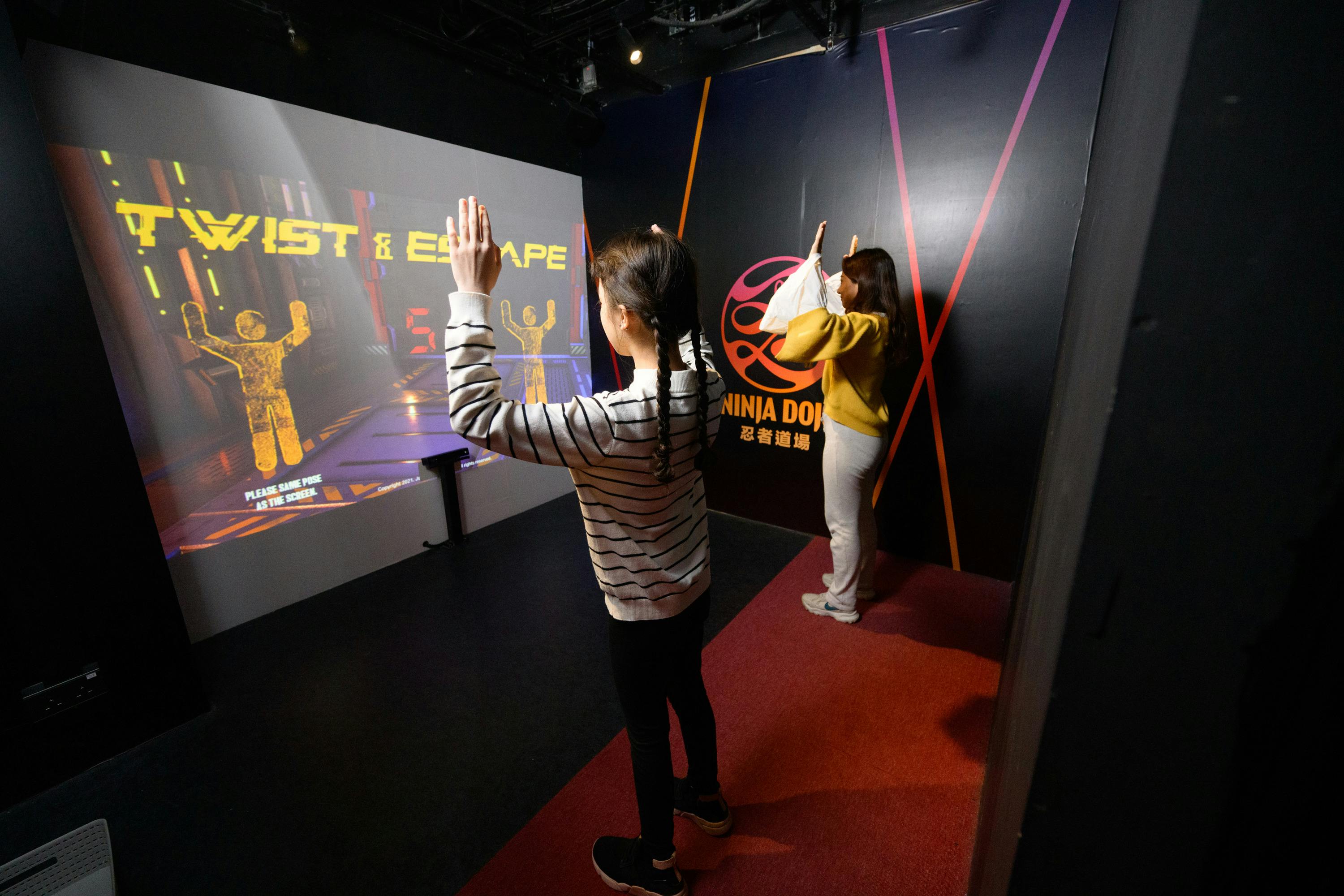 Two people interact with a virtual game on a screen in a dark room with "Ninja Dome" branding on the wall.