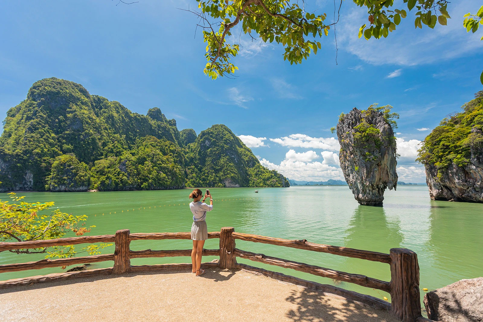 A person stands on a sandy area, photographing a tall rock formation in a green sea, surrounded by lush mountains and a clear blue sky.