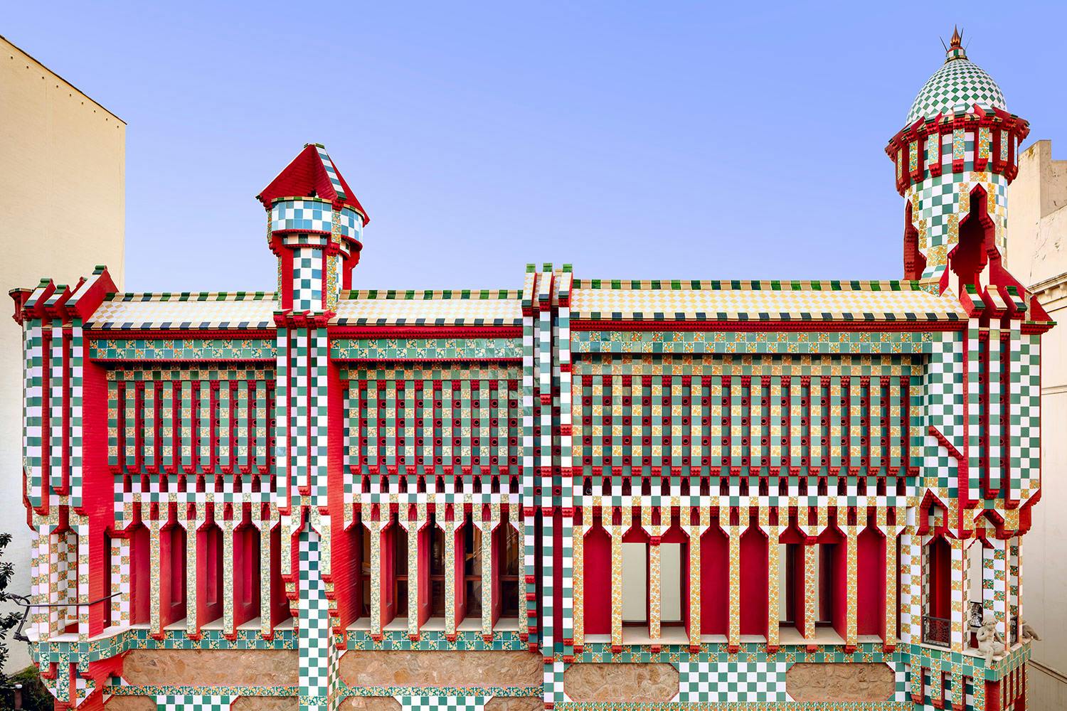Building facade with a checkered pattern in red, green, and white, featuring geometrical designs and pointed tower structures with blue sky background.