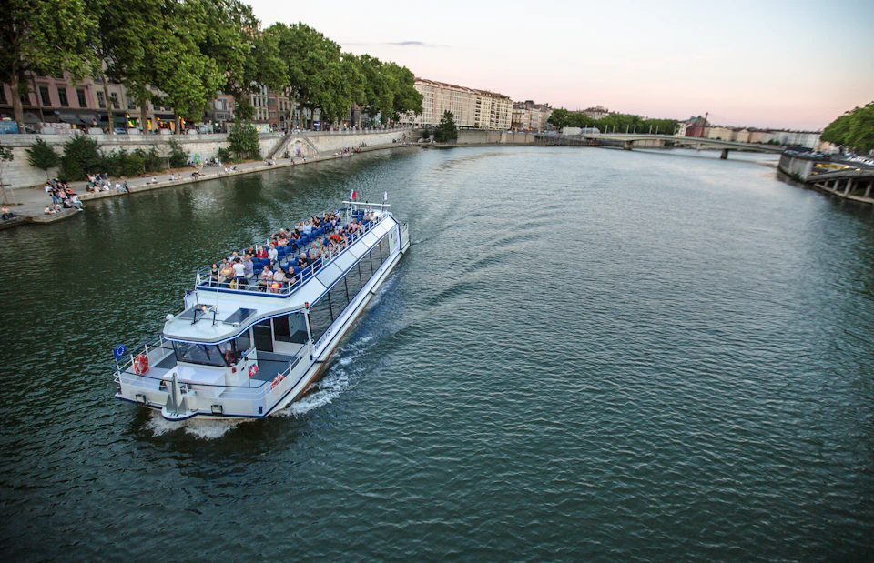 A boat with passengers sails on a river near the riverbank lined with trees and buildings.