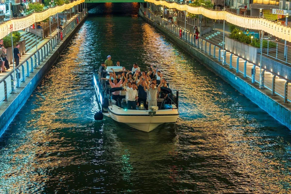 A boat filled with people raising their hands cruises down a brightly lit canal at night, with twinkling lights on the banks.