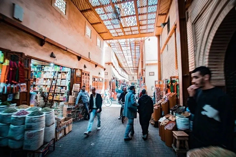 A bustling marketplace with people walking, colorful goods displayed, and sunlight filtering through a patterned roof.