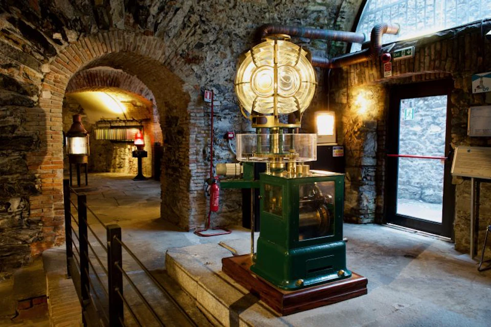 A vintage lighthouse lens displayed indoors in a historic building with stone walls and arched doorways.