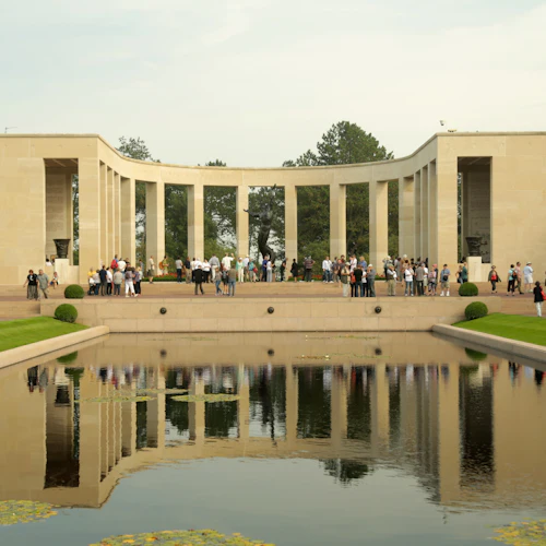 En grupp människor står framför en stor, kolonnerad minnesstruktur som speglas i en rektangulär pool. Snyggt trimmade buskar och träd omger området.