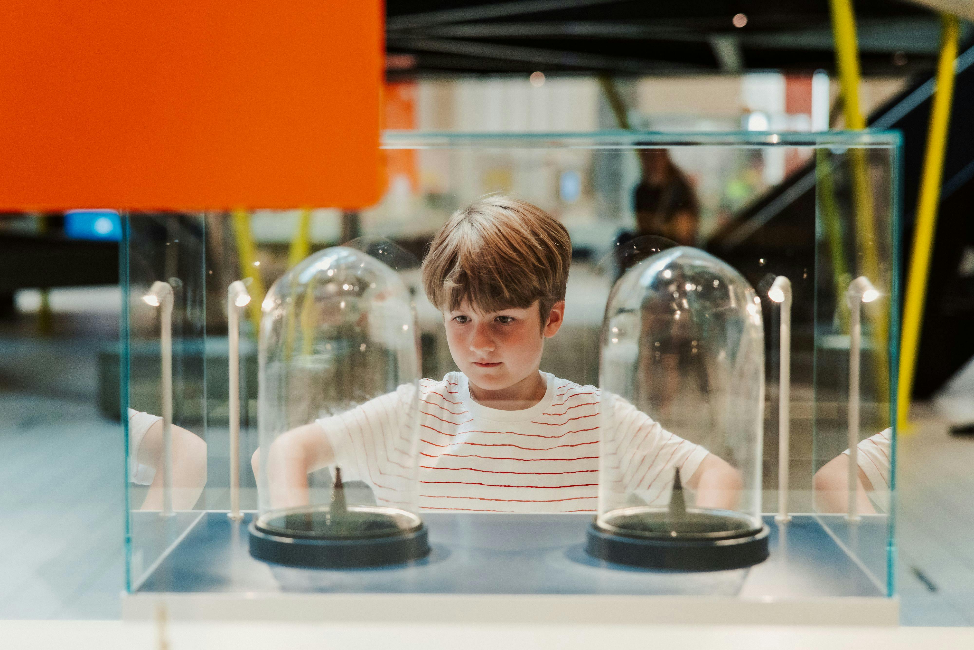 Mladý chlapec v pruhované košili upřeně hledí na dvě skleněné vystavené kopule v muzejním prostředí.
