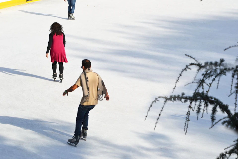 People ice skating on an outdoor rink, with one woman in a red dress and a man in a beige shirt and scarf in the foreground.