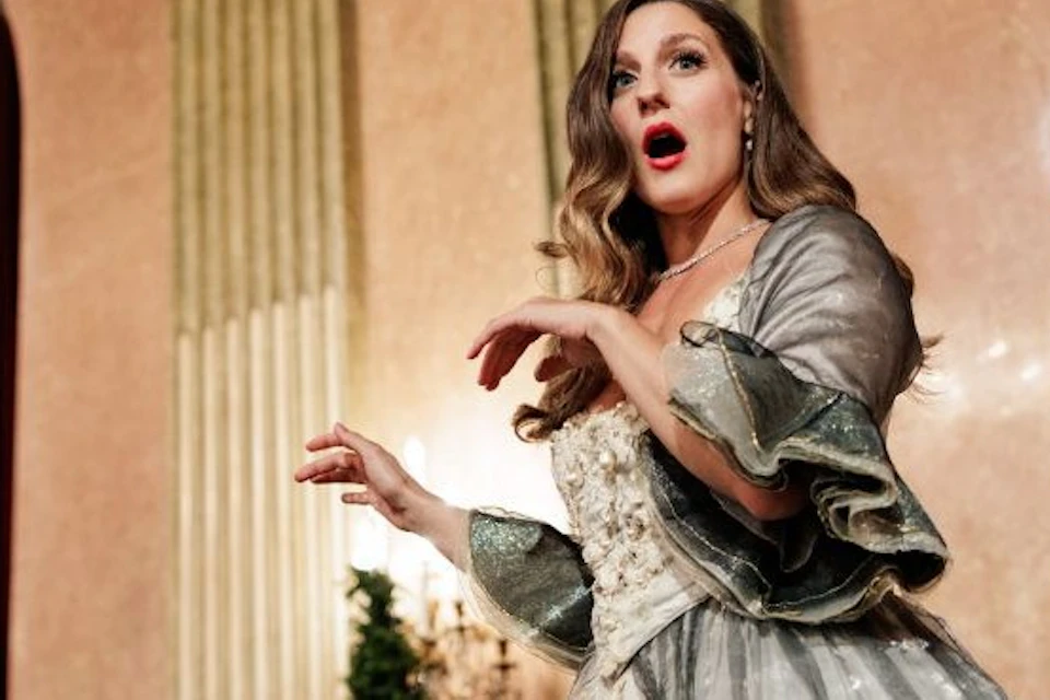 A woman in a historical-style gown performing in an ornate setting, with musicians partially visible in the background.
