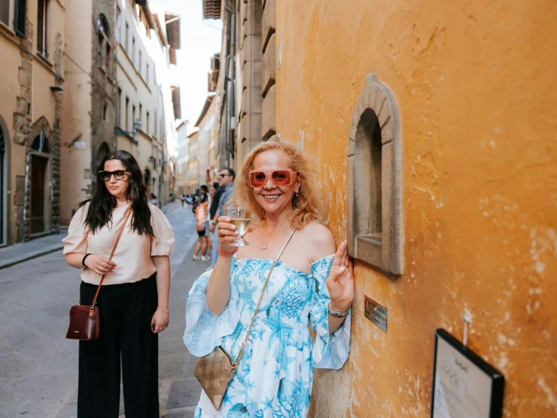 Una donna con un vestito azzurro e gli occhiali da sole sorride mentre tiene in mano un drink vicino a una piccola nicchia in una parete gialla lungo una strada.