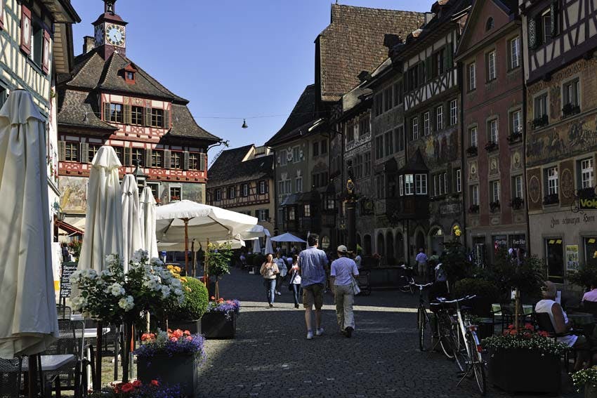 Люди гуляют по мощеной улице в европейском городе с историческими зданиями, кафе на открытом воздухе и велосипедами.